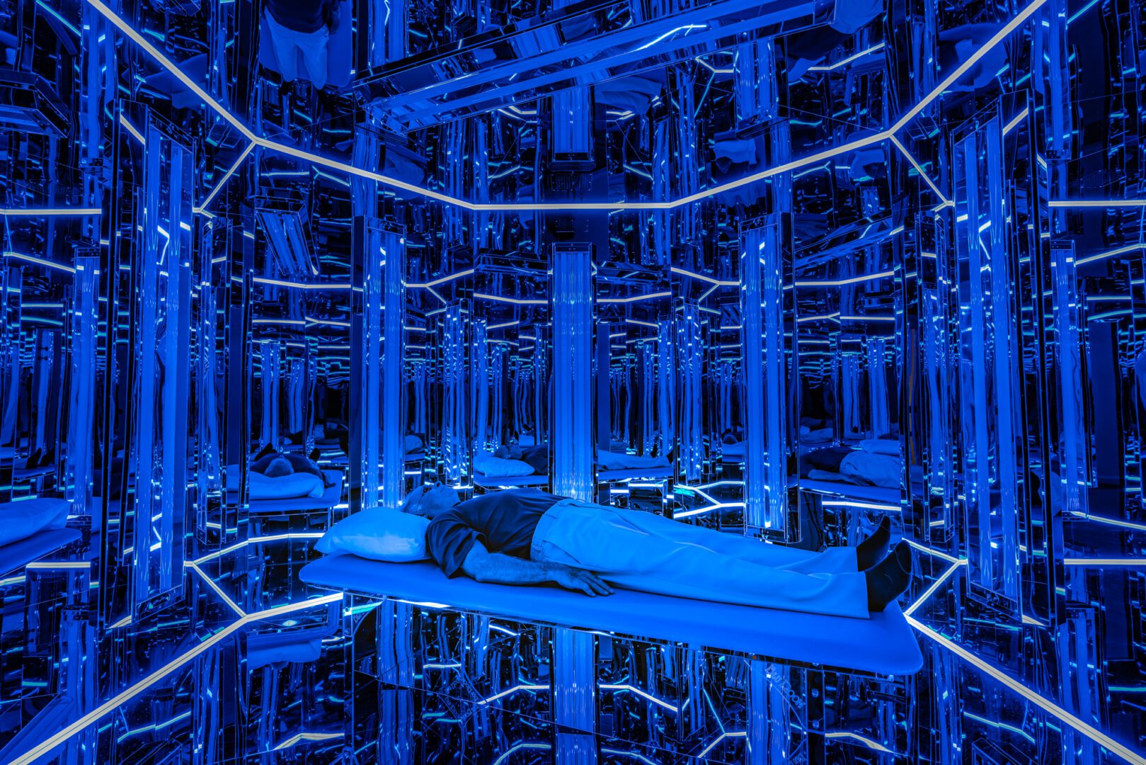 Man lies in a mirrored room lit blue.